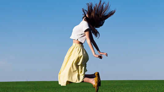 Woman in a white blouse and yellow dress jumping freely outdoors against a clear blue sky, representing joyful movement and freedom from muscle aches
