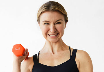 Rachel smiling in a black athletic tank top holding an orange dumbbell, representing postpartum strength and rebuilding muscle after childbirth